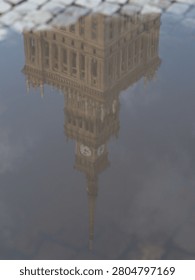 A reflection of a historic clock tower in a puddle, creating a unique perspective with cobblestone details.