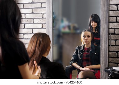 Reflection In Large Mirror Of Young Stylist Standing Behind Young Brunette Female Client Sitting In Chair In Beauty Salon