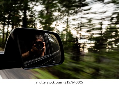 A reflection of a man holding a camera and taking photos of nature in a side mirror of a car