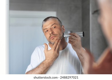 Reflection of man standing in front of the mirror and using shaver to shave his beard in bathroom in the morning