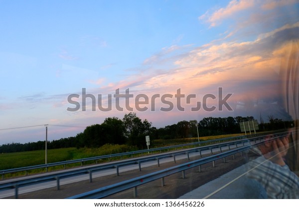 Reflection of a man in the\
window of the bus driving on the highway. Dawn or sunset outside\
the bus window. The cloudy sky and the reflection of the man.\
Tourism concept.