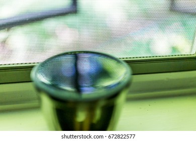 Reflection of mesh window and green foliage in cup of water or tea on windowsill