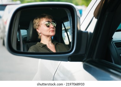 Reflection Of A Middle-aged Woman Driving A Car In The Car's Side Mirror. Woman Driving A Car. Portrait Of A Serious Woman.