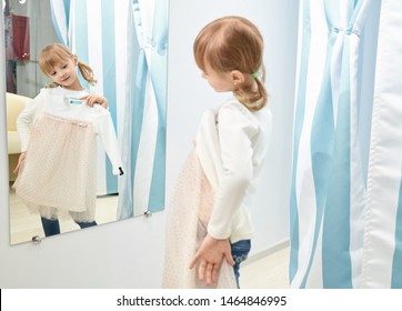 Reflection In Mirror Of Cute Girl. Little Girl Choosing And Trying Pink And White Dress In Fitting Room With Blue Curtains. Child Looking At Mirror In Mall Or Clothing Store.