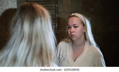 Reflection in the mirror of the face of an unhappy young blonde woman in home clothes, feeling dissatisfied with the first wrinkles, poor skin condition, ineffective skin care products - Shutterstock ID 2259158945