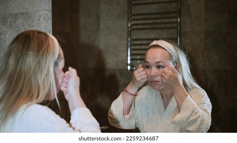 Reflection in the mirror of the face of an unhappy young blonde woman in home clothes, feeling dissatisfied with the first wrinkles, poor skin condition, ineffective skin care products - Shutterstock ID 2259234687