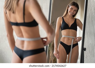 Reflection in mirror of young slim fit woman in black underwear feeling satisfied while measuring waist after dieting and workout marathon. Well-being, healthy lifestyle