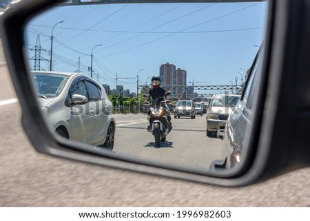 The reflection of a motorcycle in the left side mirror of a car traveling in traffic and preparing to overtake between two cars in a narrow side distance at speed. Provoking a traffic incident.