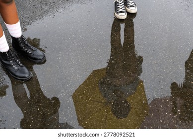 Reflection of people with umbrellas in a puddle, featuring legs in sneakers and boots on a wet pavement.