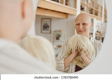 Reflection portrait of bald adult woman looking in mirror holding wig while standing in warm-toned home interior, alopecia and cancer awareness, copy space