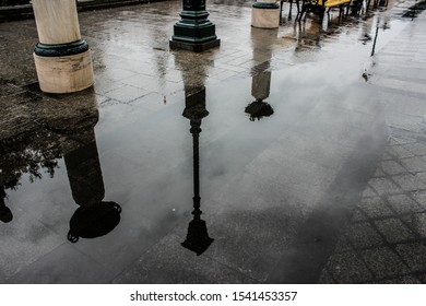 Reflection in a puddle. Columns with flower pots and street lights are visible. It's raining. The sidewalk is paved with natural stone and tiles.  The prospect goes into the distance. Autumn. Dull.