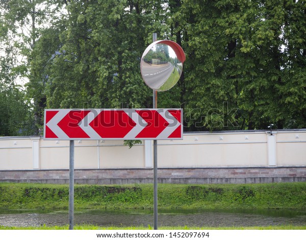 Reflection of road in the\
traffic safety mirror. Road traffic mirrow and the red traffic sign\
with two directions for driving on the forest and river background,\
soft focus