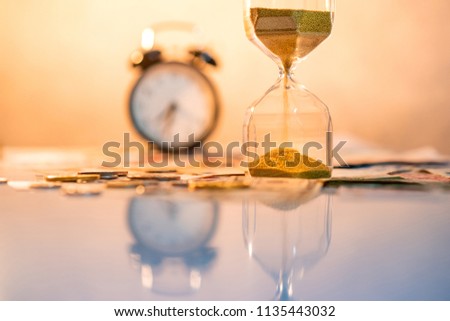 Reflection of sand running through the shape of hourglass on table with currency and clock in the background. Time investment and passing time. Urgency countdown timer for business deadline concept