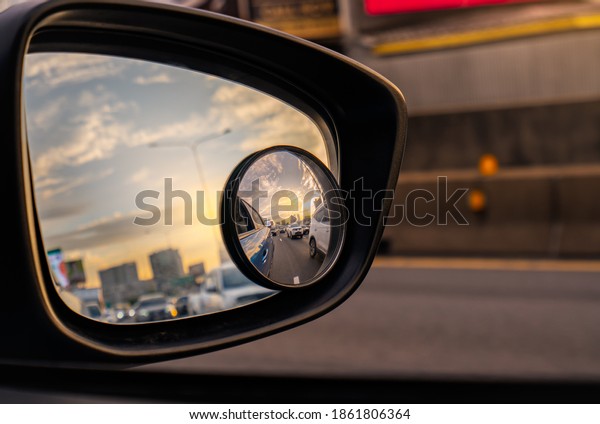 Reflection of traffic flow on asphalt road in\
side mirror of blue SUV. Car wing mirror with convex mirror for\
safety driving. View on the road and sunset sky behind cityscape\
through car side\
mirror.