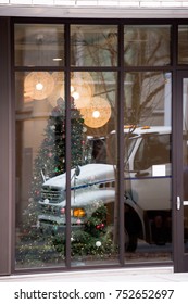 Reflection Of White Semi Truck Cab In A Large Windows Of Urban Building With A Glass Wall, Behind Which The Foredeck Room With A Decorated Christmas Tree And Glowing Large Balls Chandeliers