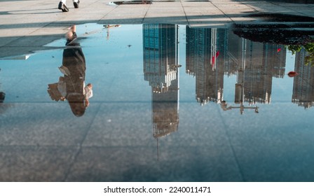 The reflection of a woman and skyscrapers in a shallow puddle on concrete ground - Powered by Shutterstock