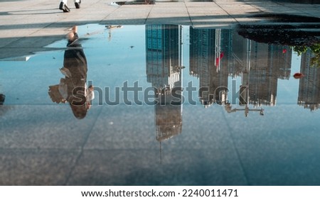 The reflection of a woman and skyscrapers in a shallow puddle on concrete ground