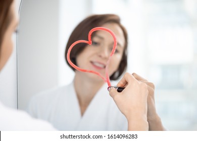 Reflection young brunette female in white bathrobe drawing heart and crimson lipstick mirror