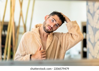 Reflection of young male in soft bathrobe touching dark hair and looking in mirror during spa session in salon - Shutterstock ID 2206579479