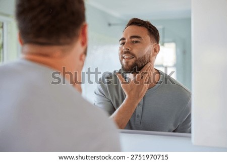 Reflection of young man touching his face while looking at mirror. Handsome man with beard examines it during grooming routine. Confident young man checking shave results with hand on face.