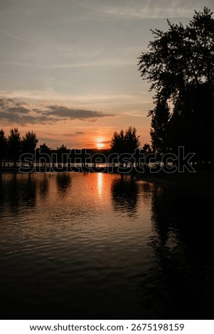 Reflections of the setting sun and trees shimmer on the calm, dark surface of the water, creating a peaceful atmosphere.