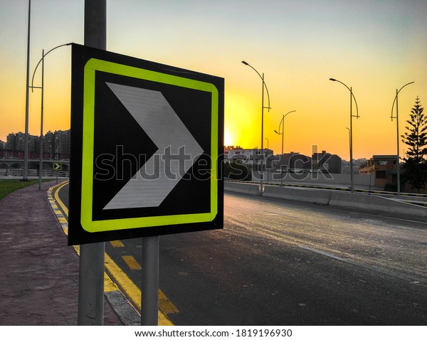 Reflective arrow road sign to warn of a sharp\
turn with sunset in the\
background