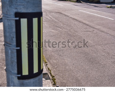 A reflective caution marker is strapped to a concrete utility pole situated next to an asphalt road. The high-visibility yellow and black striped strip is designed for night safety on a residential street corner.