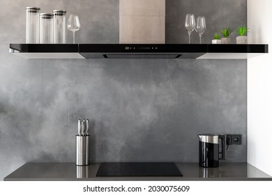 Reflective countertop with built-in induction cooker, exhaust hood with touch controls and glasses on top of it, set of knives and black kettle in modern kitchen in contemporary appartment