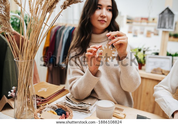 Reflective woman holding\
wooden car pendant shaped as a snowflake, lost in a thought,\
looking to the side. She\'s touching one of the spikes. Leaning\
elbow on the table.