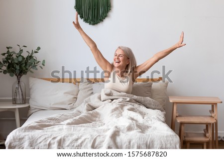 Refreshed elderly 50s female wakes up in morning stretches seated in bed in light bedroom at home, middle-aged woman feels happy and peppy after enough sleeping, greeting new day, good morning concept