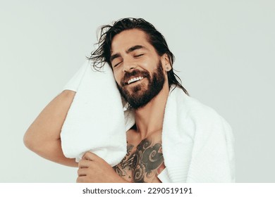 Refreshed young man wiping his hair with a bath towel after taking a relaxing shower. With a smile on his face, a handsome caucasian man enjoys his grooming and self-care routine in a studio. - Powered by Shutterstock