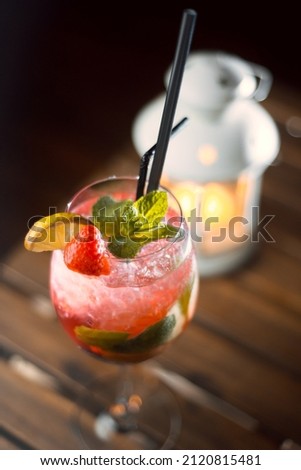Refreshing alcoholic cocktail with mint, strawberry and lime. A candle burns on a wooden table and in the background
