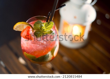 Refreshing alcoholic cocktail with mint, strawberry and lime. A candle burns on a wooden table and in the background