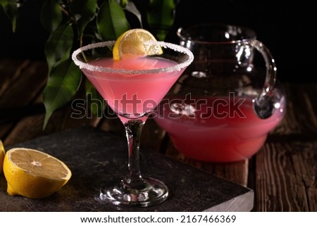 Refreshing citrus cocktail with sugar rim and lemon, on a stone slab and wooden table.  In the background, a decanter with a cocktail and leaves of a tree, at night