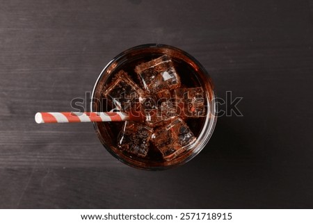 Refreshing cola with ice cubes and drinking straw in glass on black wooden table, top view