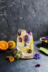 Refreshing Cold Drink With Lemon, Orange, Cucumber And Viola Flowers In Two Glasses. Fruit Cocktail With Ice And Edible Flowers.