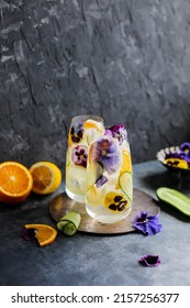 Refreshing cold drink with lemon, orange, cucumber and viola flowers in two glasses. Fruit cocktail with ice and edible flowers.