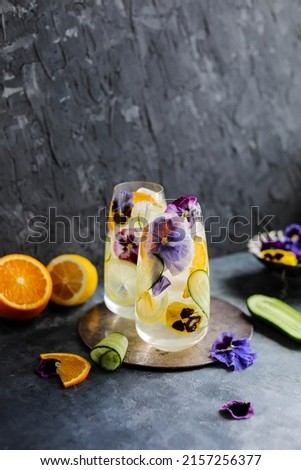 Refreshing cold drink with lemon, orange, cucumber and viola flowers in two glasses. Fruit cocktail with ice and edible flowers.