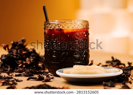 Refreshing cold hibiscus tea in a patterned glass with a black straw and dried herbal leaves scattered on a rustic wooden table surface nearby