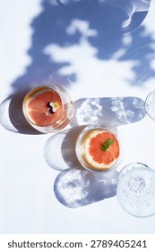 Refreshing grapefruit drinks in glasses with mint and edible flower on a bright white surface with strong afternoon sun shadows.
