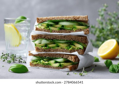 A refreshing and healthy stack of avocado and cucumber sandwiches on whole wheat bread, garnished with fresh basil and served with a glass of infused water.