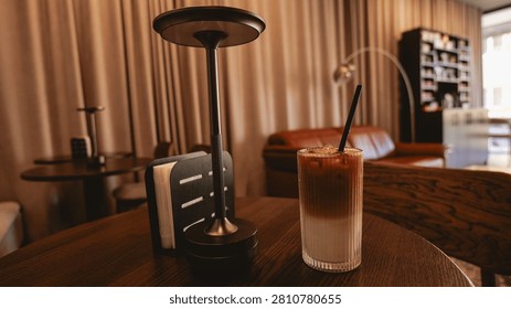 A refreshing iced coffee with distinct layers of milk and espresso sits on a wooden table next to a modern lamp and a napkin holder