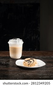 A refreshing iced coffee in a plastic cup with condensation, accompanied by a delectable chocolate chip cookie on a white plate, resting on a dark wooden table in a cafe