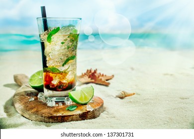 Refreshing Iced Rum, Lime And Mint Mojito Cocktail Standing In A Tall Glass On An Old Wooden Board On The Golden Sand Of An Idyllic Tropical Beach To Celebrate A Summer Vacation