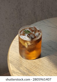 Refreshing iced tea served in a clear glass with ice cubes and fresh rosemary garnish on a rustic wooden table. Warm natural sunlight creates a vibrant amber glow, making this beverage ideal for café menus, summer drink promotions, healthy lifestyle concepts, and commercial food photography.