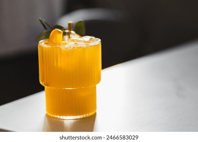 A refreshing orange cocktail, garnished with a slice of orange and green leaves, served in a textured glass on a reflective surface. The soft lighting in the background adds to its appeal - Powered by Shutterstock