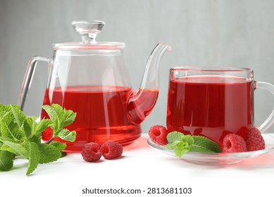 Refreshing raspberry tea in glass cup, teapot, fresh berries and mint leaves on white wooden table against grey background, closeup