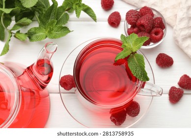 Refreshing raspberry tea in glass cup, teapot, fresh berries and mint leaves on white wooden table, flat lay
