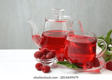 Refreshing raspberry tea in glass cup, teapot, fresh berries and mint leaves on white wooden table against grey background, closeup. Space for text