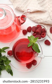 Refreshing raspberry tea in glass cup, teapot, fresh berries and mint leaves on white wooden table, flat lay